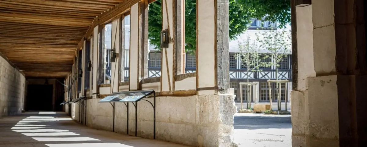 A picturesque view of the medieval courtyard of Aître Saint-Maclou, showcasing its timber-framed architecture with large windows and wooden beams, leading to an open square with more half-timbered buildings and lush greenery.