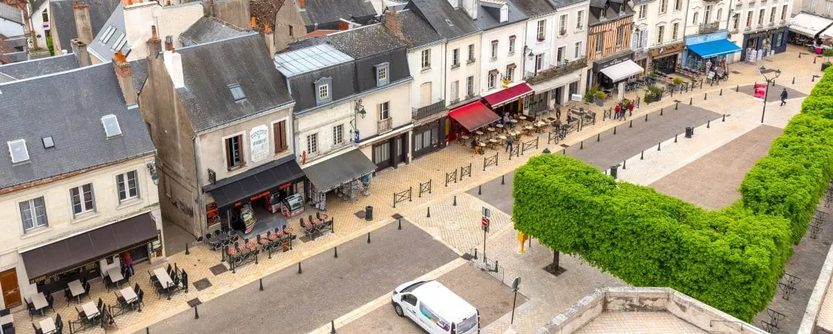 An aerial view of a quaint town center featuring rows of charming, old-style buildings with storefronts, cafes, and outdoor seating. The scene includes a white van parked on the cobblestone street and well-maintained greenery.