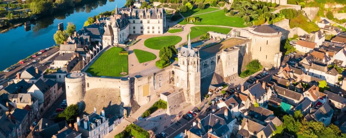 Aerial view of Amboise town featuring a historic castle with stone walls, surrounded by lush green lawns, residential buildings, and a river running through the town.