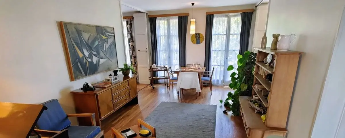 A cozy living and dining area in Apartment Perret, Le Havre, featuring wooden furniture, a large abstract painting, and floor-to-ceiling windows with curtains.