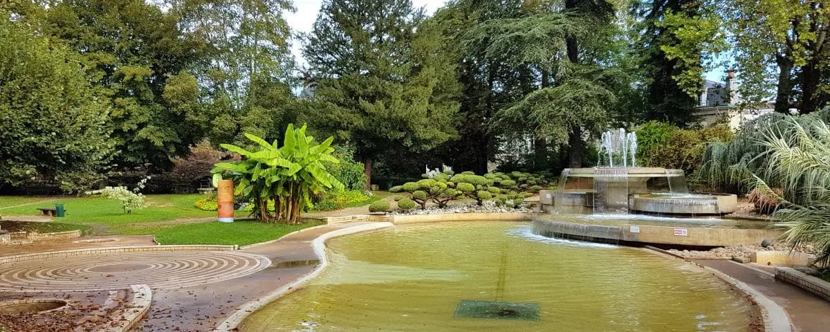 A serene garden in Périgueux featuring a circular fountain with water jets, surrounded by lush greenery and manicured shrubs. The setting includes a winding path and tall trees in the background, offering a peaceful atmosphere.