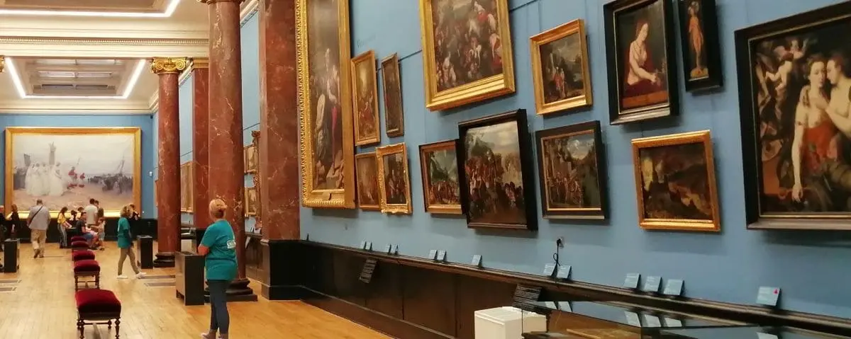 A large hall in a museum with blue walls adorned with framed paintings and sculptures. People are observing the artworks, and there are red cushioned seats along the wooden floor.