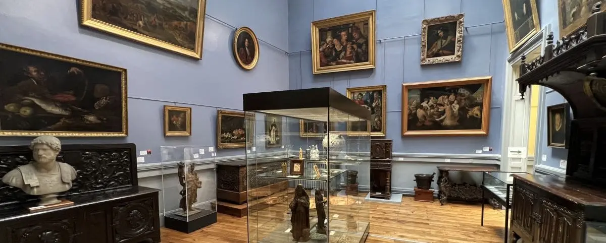 An interior view of a museum room showcasing various artworks and artifacts. The walls are adorned with framed paintings, and a central glass display case holds sculptures and historical pieces. The room's classical decor includes busts and wooden furniture.
