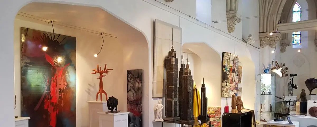 Interior of an art gallery within a chapel, featuring modern sculptures and paintings displayed against white walls and arched ceilings.