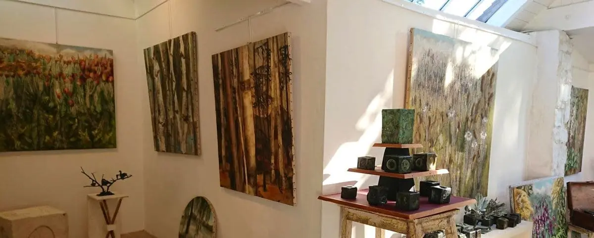 An art gallery displaying various paintings, including landscapes and abstract works. The room is well-lit with natural light streaming in from a skylight, and there are also ceramic pieces arranged on a small table.