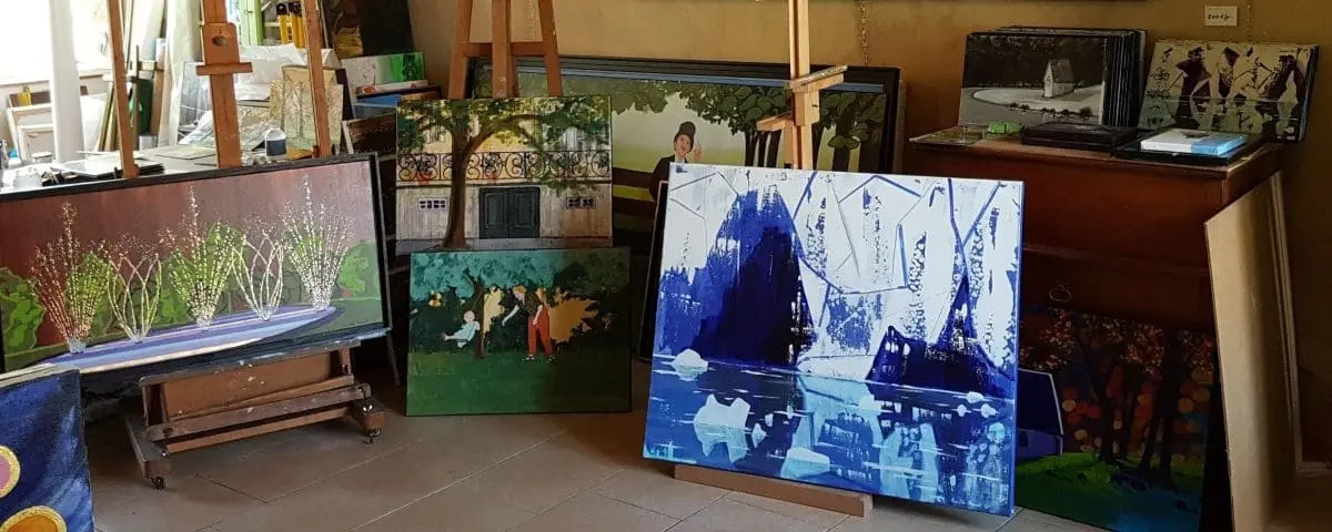A vibrant artist's workshop with various paintings displayed on easels and propped against the walls. The artworks include a mix of colorful landscapes, abstract scenes, and garden views.