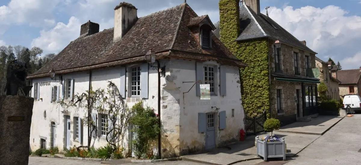 A charming, rustic building with white walls and a steeply pitched roof in the village of Saint-Céneri-le-Gérei. The building is adorned with ivy and surrounded by a quaint street and flower boxes.