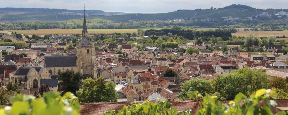 A scenic view of the town of Ay, featuring a mix of traditional houses and a prominent church steeple in the center. The town is surrounded by lush green fields and rolling hills under a partly cloudy sky, with vineyards in the foreground.