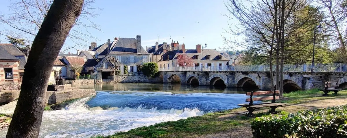 Serene scene in Azay-le-Rideau with a calm river, a quaint stone bridge, and traditional French houses in the background, framed by trees and a park bench in the foreground.