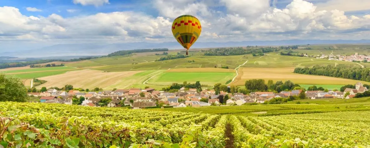 A colorful hot air balloon floating above vast, scenic vineyards with a small village in the distance under a partly cloudy sky.