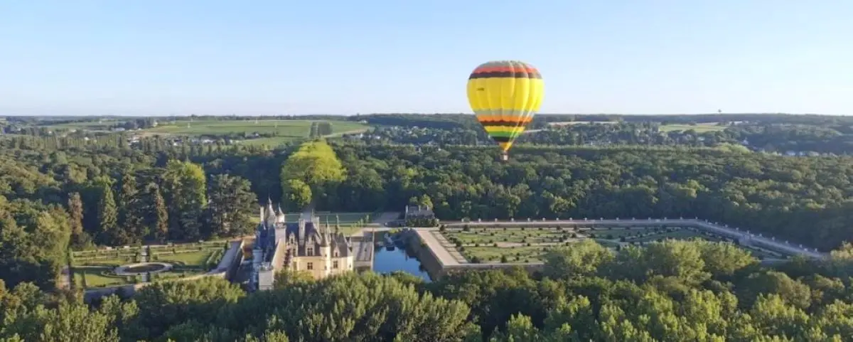 A colorful hot air balloon floating over the lush green landscape of the Loire Valley, with a historic chateau and extensive gardens below.
