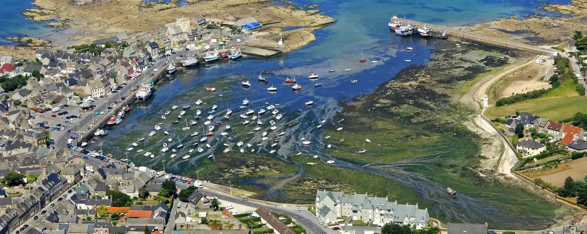 Aerial view of Barfleur village with a harbor filled with boats, rocky shorelines, and a mix of residential and commercial buildings along the waterfront.
