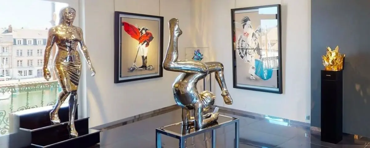 A view inside the Bartoux Gallery in Honfleur, showcasing modern art pieces including metallic sculptures and vibrant paintings. The room is well-lit, highlighting the intricate details of the artwork.