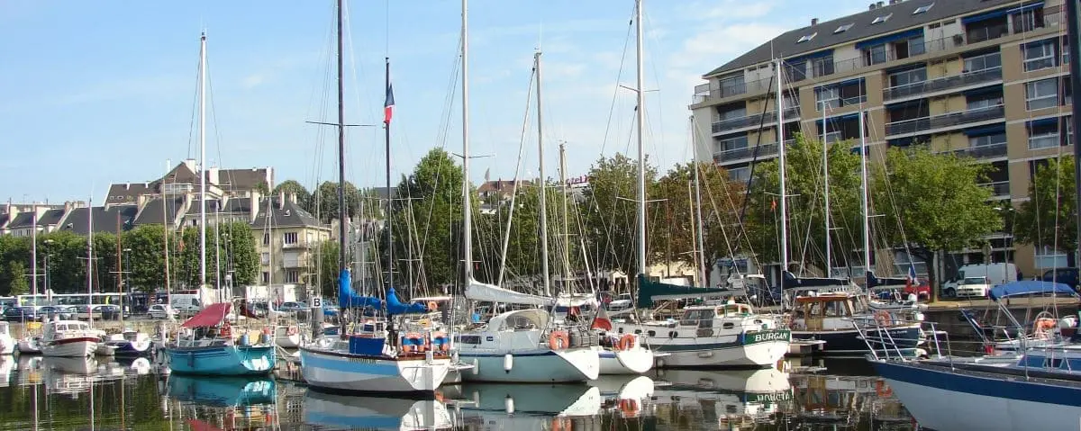 Boats docked at the marina in Caen, with apartment buildings and trees in the background. The clear sky and calm water reflect the peaceful ambiance of the marina.