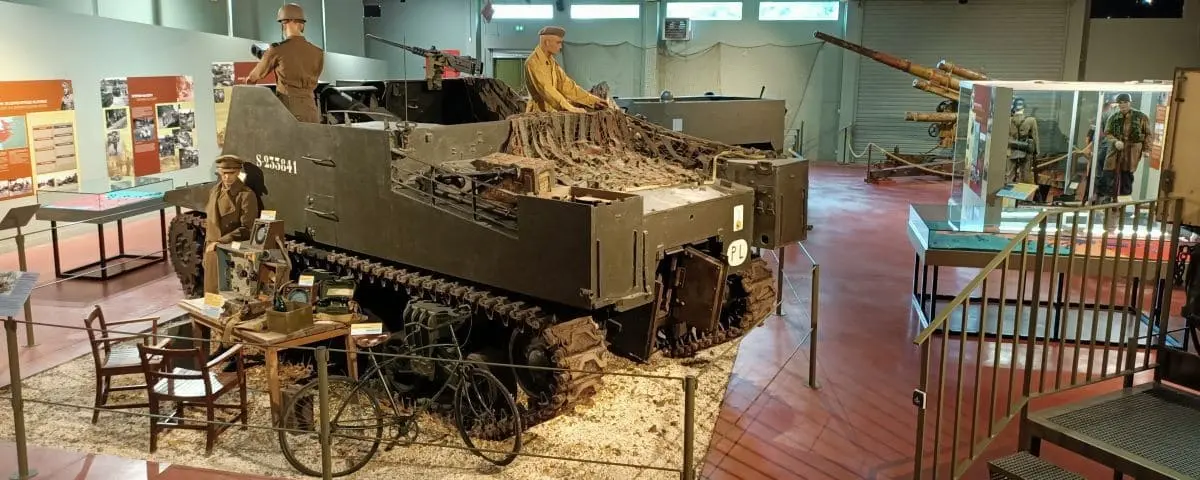 A military museum exhibit featuring a large armored vehicle with mannequins dressed in military uniforms, surrounded by various wartime artifacts and displays.