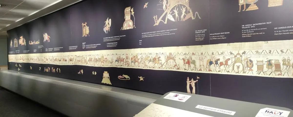 A long, detailed segment of the Bayeux Tapestry displayed in a museum, depicting historical scenes with descriptive text above.