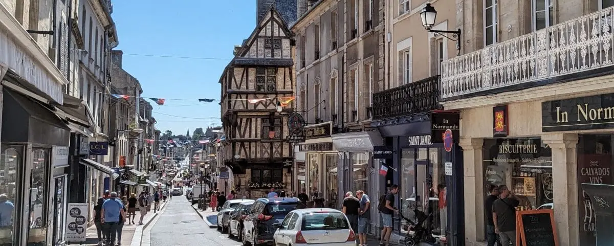 A bustling street in the center of Bayeux, lined with shops and half-timbered buildings, with people walking and cars parked along the road.