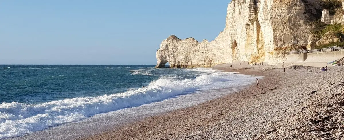 A serene beach in Etretat with pebbly shores, gentle waves, and dramatic white cliffs under a clear blue sky.