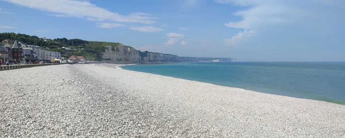A wide pebble beach in Fécamp, France, with clear blue skies and calm turquoise waters. The beach is bordered by cliffs and a quaint seaside town.
