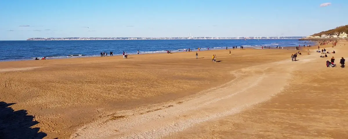 A wide, sandy beach in Trouville stretching to the horizon, with people scattered along the shore and the calm sea in the background under a clear sky.