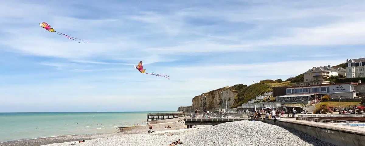 A scenic beach view with pebbles, calm sea, and colorful kites flying in the blue sky. Cliffs and seaside buildings are visible in the background.
