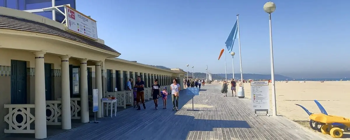 People stroll along the wooden boardwalk at Les Planches in Deauville, adjacent to the sandy beach and lined with beach huts.