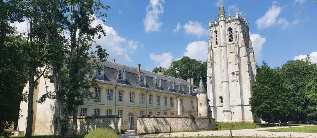 A sunny day view of the Bec Hellouin Abbey with its historical stone buildings and tall tower, surrounded by lush greenery and a bright blue sky with scattered clouds.