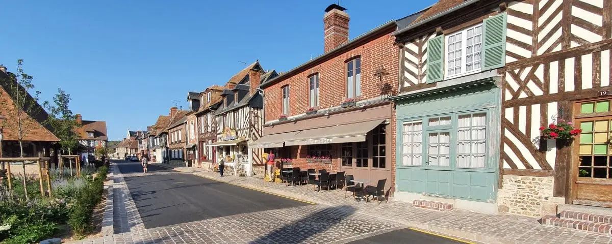Quaint street in Beuvron-en-Auge village lined with half-timbered houses and a few outdoor café tables under a clear blue sky.
