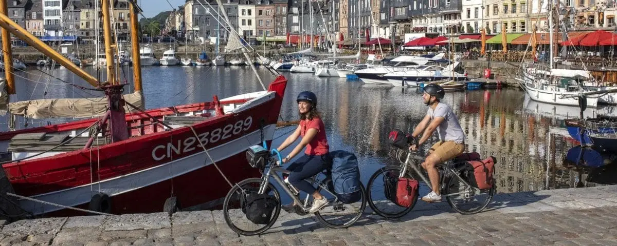 Two cyclists, a man and a woman, ride their bikes along the waterfront in Honfleur. They are passing a red boat docked in the harbor, with a picturesque backdrop of colorful buildings and other boats.