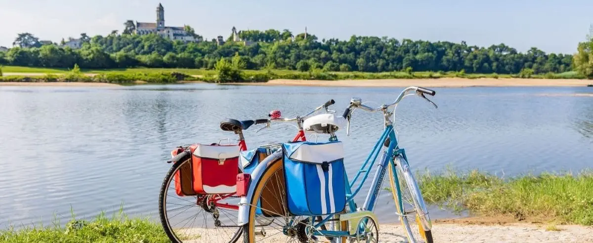 Two bicycles with panniers parked by a river in the Loire Valley, with a scenic view of a castle and dense greenery in the background.