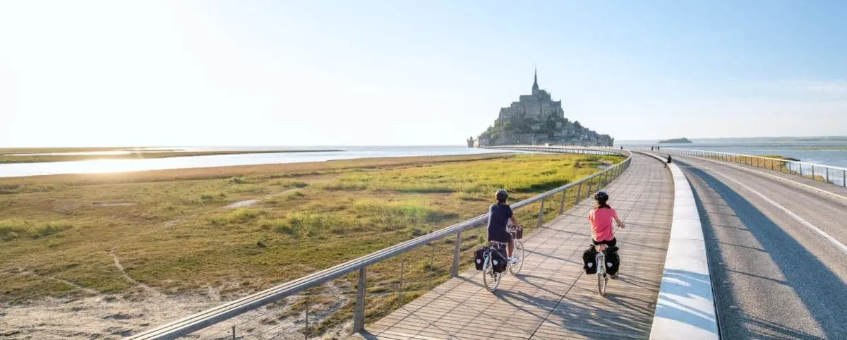 Two cyclists ride along a wooden path towards Mont Saint-Michel, with a clear sky and scenic views of the surrounding bay and grasslands.