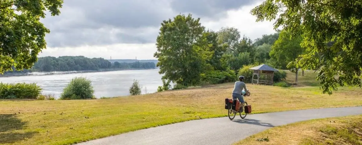 A person riding a bicycle along a paved path beside a river, surrounded by greenery. The sky is cloudy, and there is a small wooden structure visible in the distance.