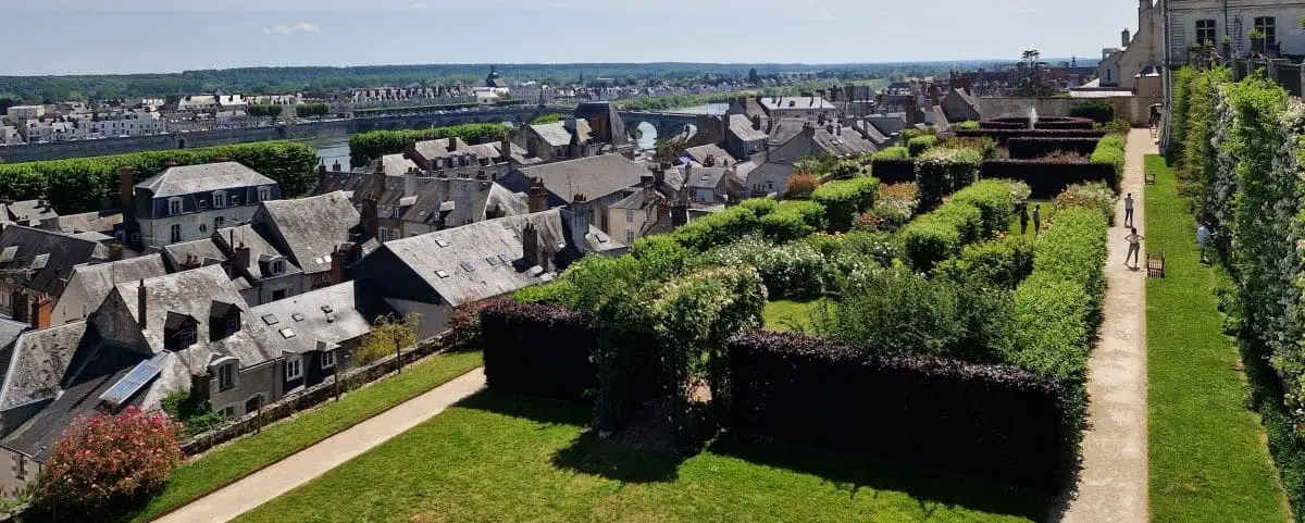 Overlooking a picturesque town with a river in the distance, featuring well-maintained gardens with hedges and pathways.