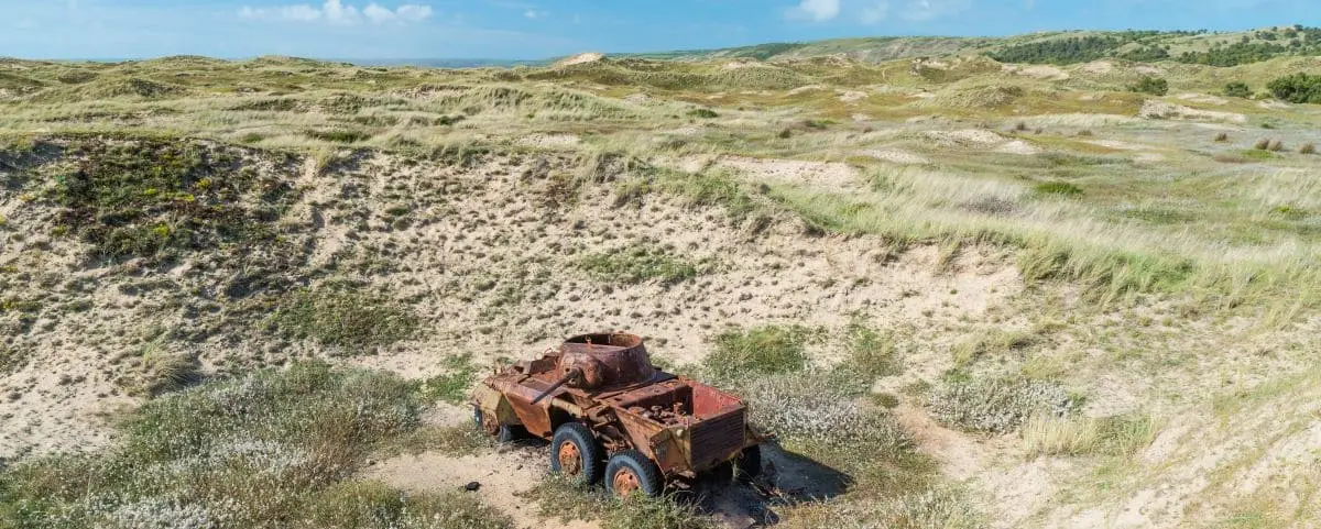 Expansive sandy dunes at Biville with sparse vegetation and a rusty old vehicle partially buried in the sand under a bright, sunny sky.