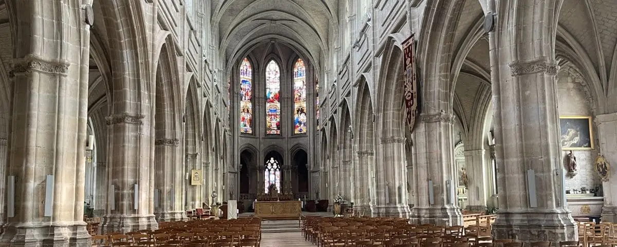 The interior of a majestic cathedral with tall, arched ceilings, stained glass windows, and rows of wooden chairs.