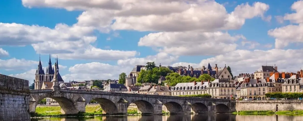 Picturesque view of Blois town featuring a stone bridge over a river, historic buildings, and a cathedral spire against a backdrop of a partly cloudy sky.