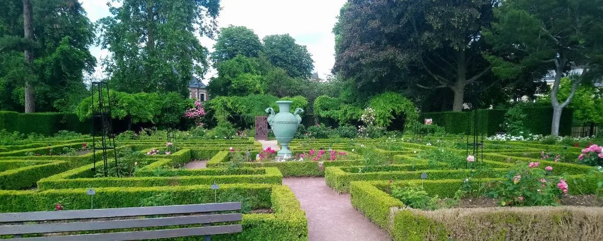 A well-manicured garden in the Botanical Garden of Rouen, with neatly trimmed hedges forming a maze-like pattern, vibrant flowers, and a large, decorative urn at the center.