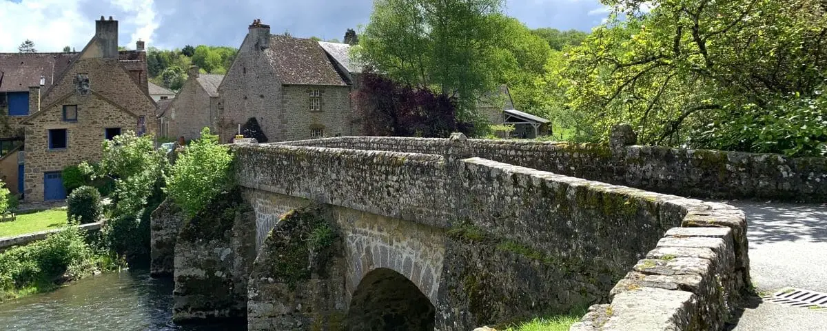 A picturesque stone bridge crossing over a serene river, surrounded by lush greenery and historic stone buildings in Saint-Céneri-le-Gérei.