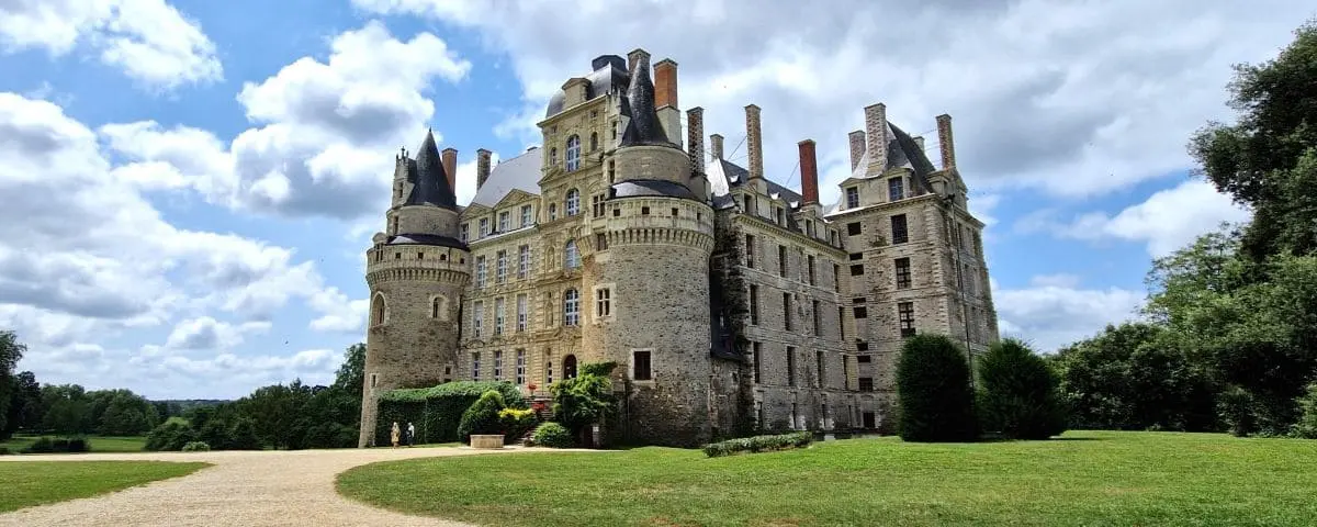 An imposing, historic castle with tall, round towers and numerous chimneys, surrounded by a manicured lawn and a clear sky.