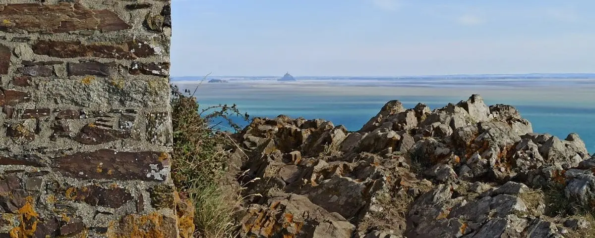 A rocky coastline with a stone structure on the left, overlooking a distant islet across a vast body of water. The scene is serene and expansive.