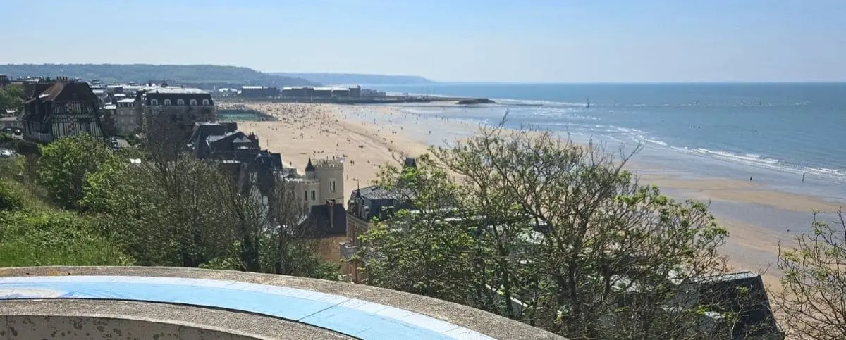 A panoramic view of Trouville's coastline, showing a beach with many people, lush greenery, and historic buildings with the sea stretching into the distance under a clear blue sky.