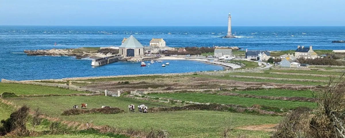 Scenic coastal view of Cap de la Hague with traditional stone buildings, a lighthouse in the distance, and green fields leading to the blue sea.