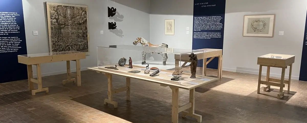 A room in the Carroi Museum in Chinon featuring various artifacts displayed on wooden tables with glass covers. There are sculptures, a model horse, and wall-mounted artworks in the background.