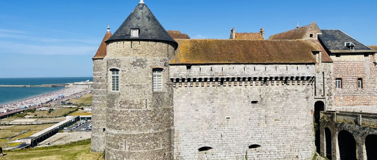 The historic Dieppe Castle, featuring large stone walls and a round turret, with the coastline and beach visible in the background under a clear sky.
