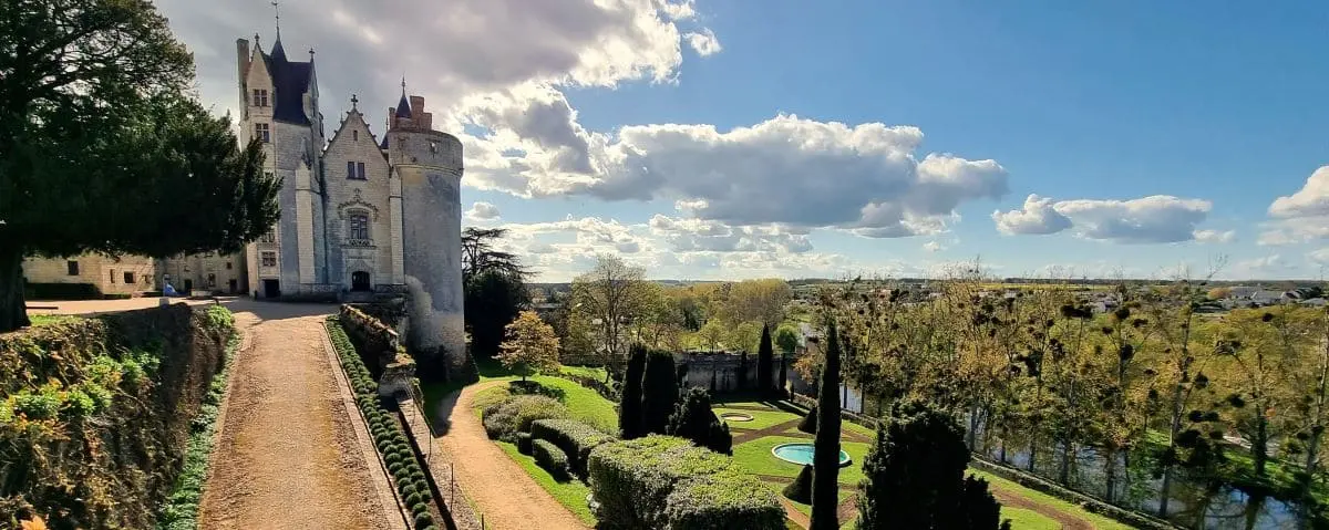 The impressive Castle of Montreuil-Bellay with its fortified walls, towers, and well-manicured gardens, under a partly cloudy sky.