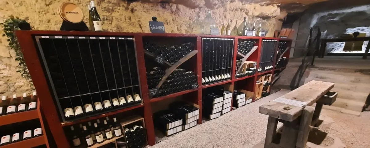 A rustic wine cellar with several wooden shelves filled with wine bottles, neatly organized. The room has a cozy, historic atmosphere with stone walls and a wooden bench in the foreground.