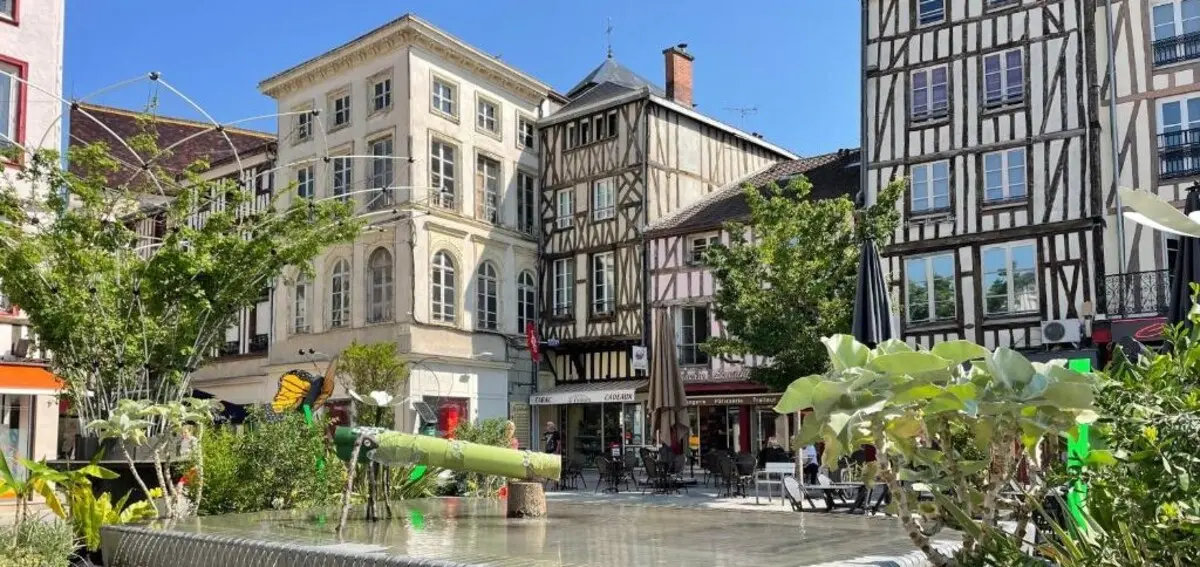 A picturesque town square with half-timbered houses, vibrant shops, and cafes. A small garden with plants and a playful water feature in the foreground.