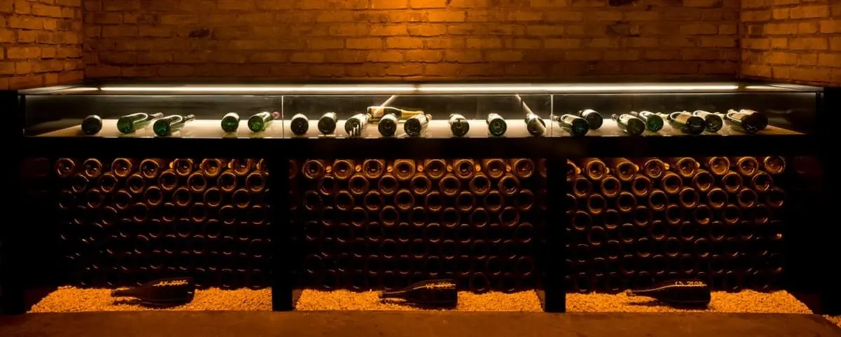A dimly lit cellar with rows of champagne bottles stored horizontally on shelves against a brick wall. The bottles are arranged in neat rows, creating an organized and atmospheric setting.
