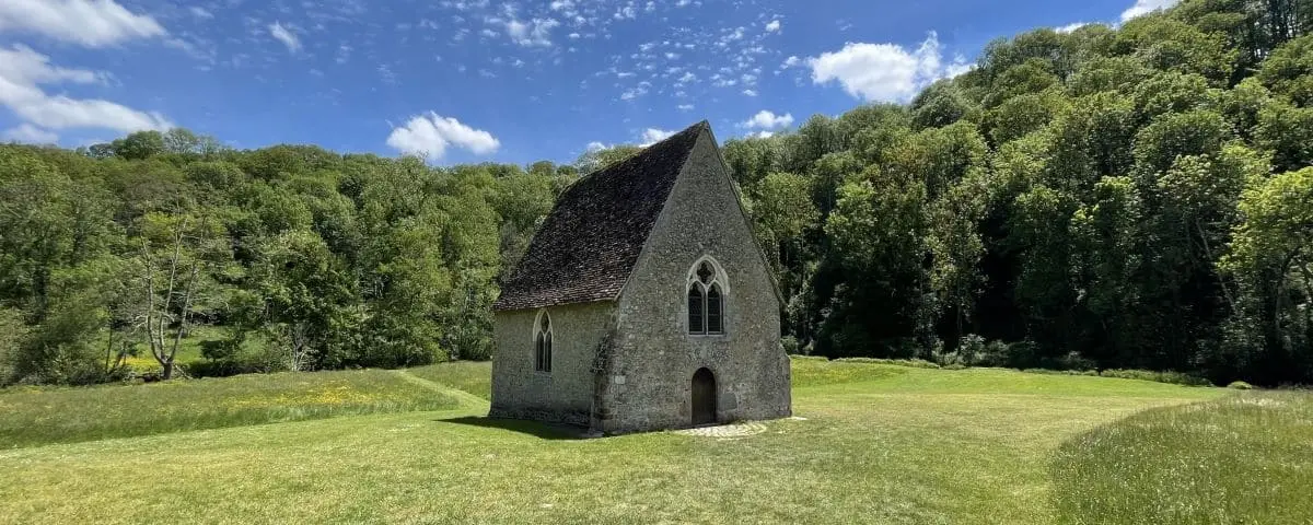 A small, stone chapel with a steep roof, set in a tranquil grassy area bordered by dense forest. The chapel stands alone, offering a serene and peaceful atmosphere.