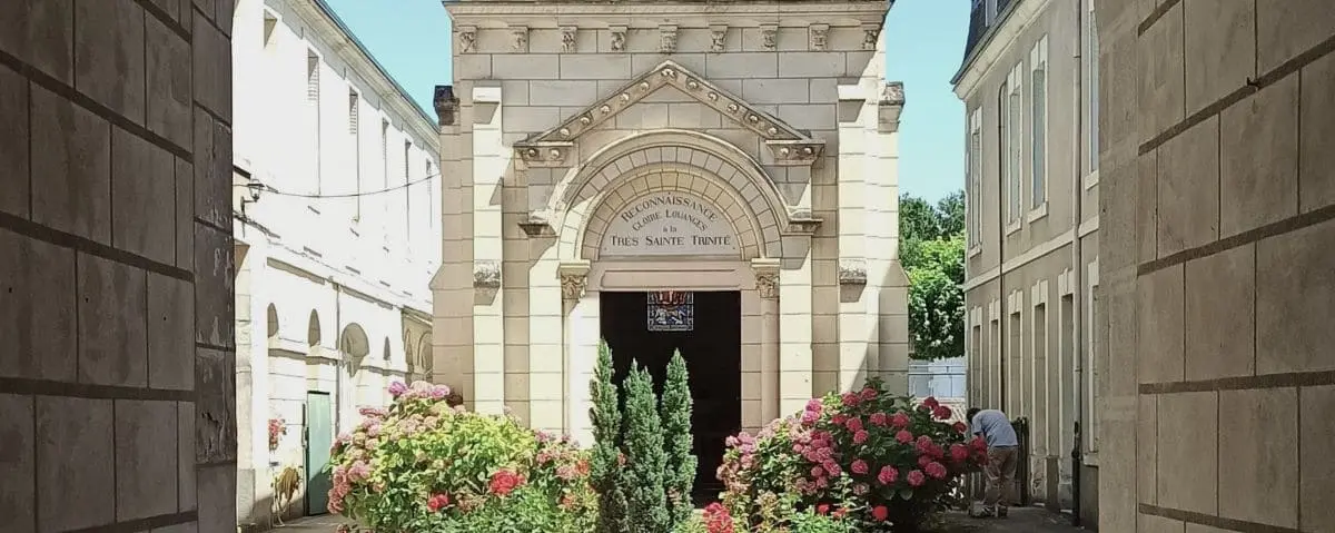 A charming chapel in Richelieu with a stone facade, surrounded by blooming flowers and greenery, nestled between two buildings.